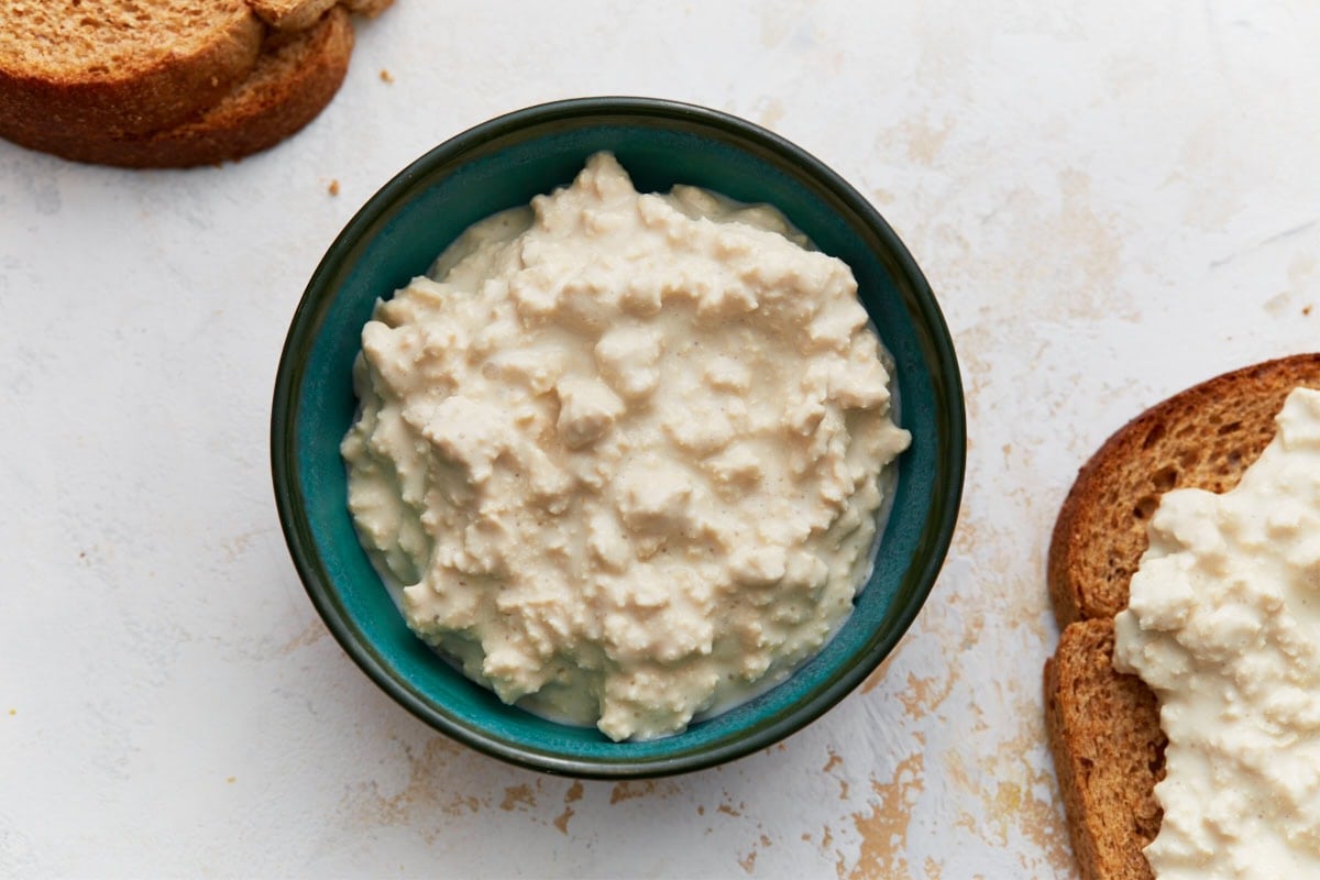 A teal bowl of finished dairy-free cottage cheese next to slices of toasted bread on a light surface.