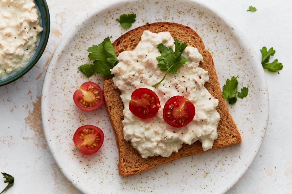 A slice of toast topped with dairy-free cottage cheese, cherry tomato halves, and fresh parsley on a white plate.