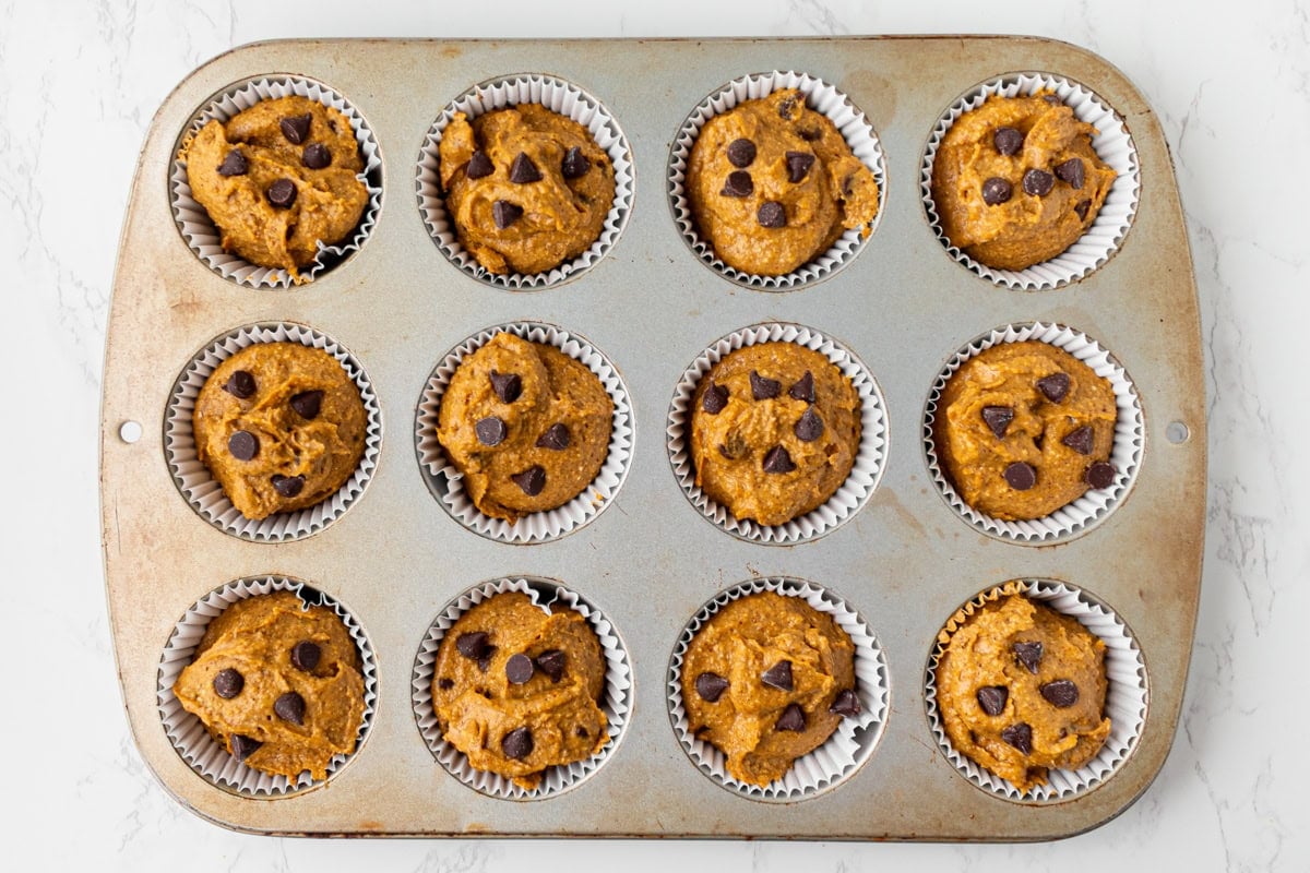 Muffin tin lined with paper cups filled with pumpkin batter and topped with chocolate chips, ready for the oven.