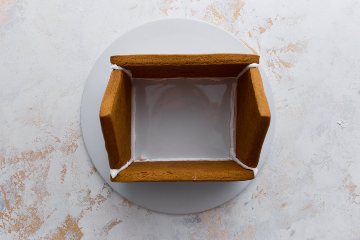 The base of the gingerbread house being assembled on a round platter, with four cookie walls secured together using white royal icing.