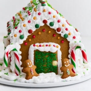 A fully assembled gluten-free gingerbread house decorated with white royal icing, colorful candies, candy canes, and two small gingerbread people by the doorway.
