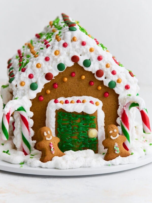 A fully assembled gluten-free gingerbread house decorated with white royal icing, colorful candies, candy canes, and two small gingerbread people by the doorway.
