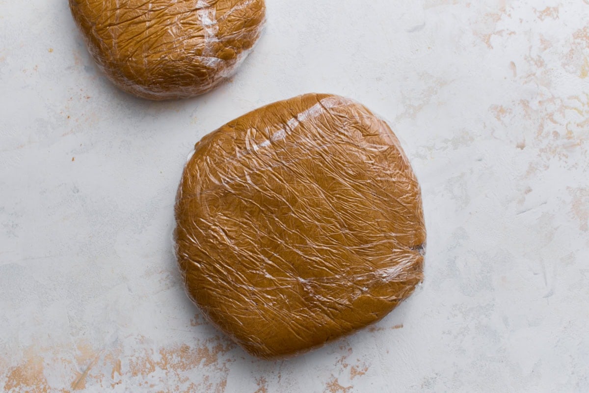 Two flattened discs of gingerbread dough wrapped tightly in plastic wrap, resting on a light surface.