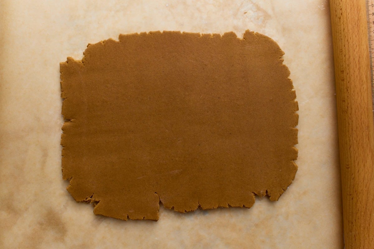 A sheet of rolled-out gingerbread dough between parchment paper, about ¼ inch thick, ready for cutting house templates.