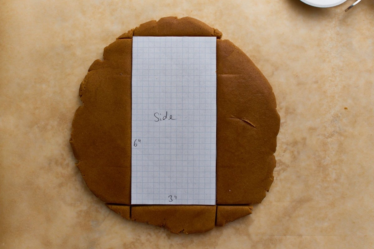 Rolled-out gingerbread dough with a rectangular graph-paper template labeled “Side” placed on top, showing measurements for cutting the house wall.