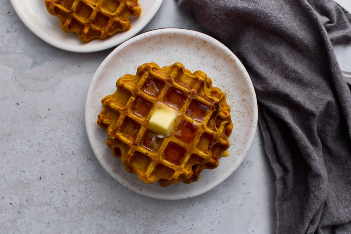 Stacked pumpkin waffle topped with butter and drizzled with syrup on a plate.