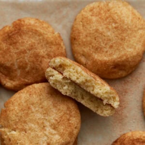 Soft gluten-free snickerdoodle cookies on parchment paper, with one cookie broken in half to show the fluffy, tender inside and cinnamon sugar coating.