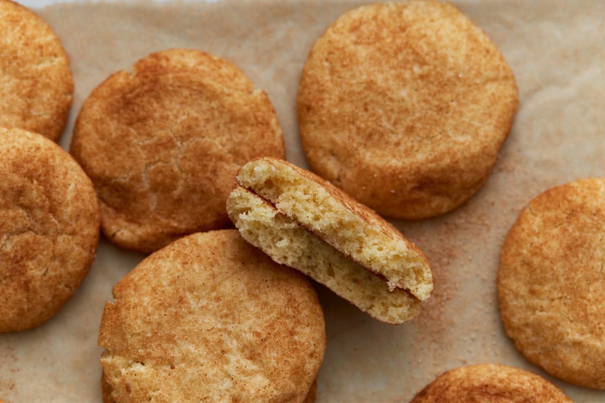 Soft gluten-free snickerdoodle cookies on parchment paper, with one cookie broken in half to show the fluffy, tender inside and cinnamon sugar coating.
