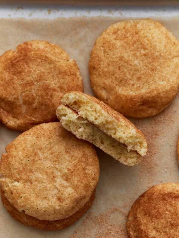 A batch of soft gluten-free snickerdoodle cookies on parchment paper, with one cookie broken in half to show the fluffy, cinnamon-speckled center.