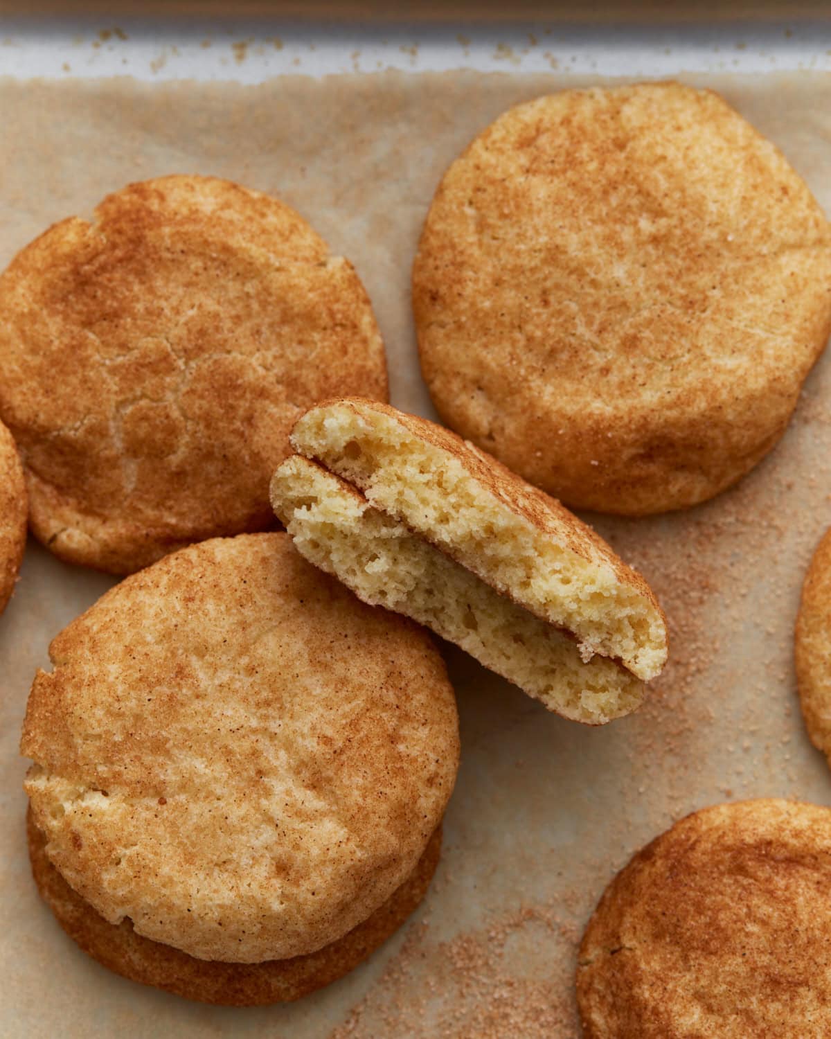 A batch of soft gluten-free snickerdoodle cookies on parchment paper, with one cookie broken in half to show the fluffy, cinnamon-speckled center.