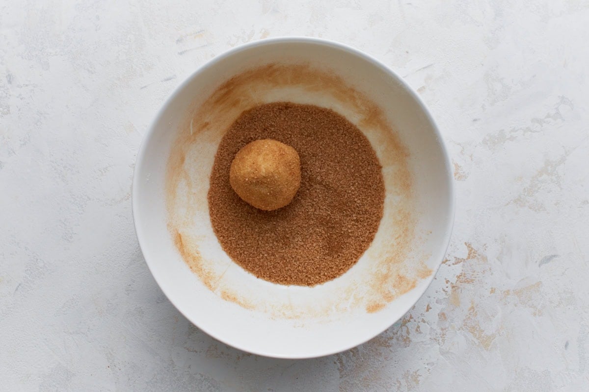 A cookie dough ball coated in a mixture of cinnamon and sugar in a white bowl, showing the classic snickerdoodle coating step.