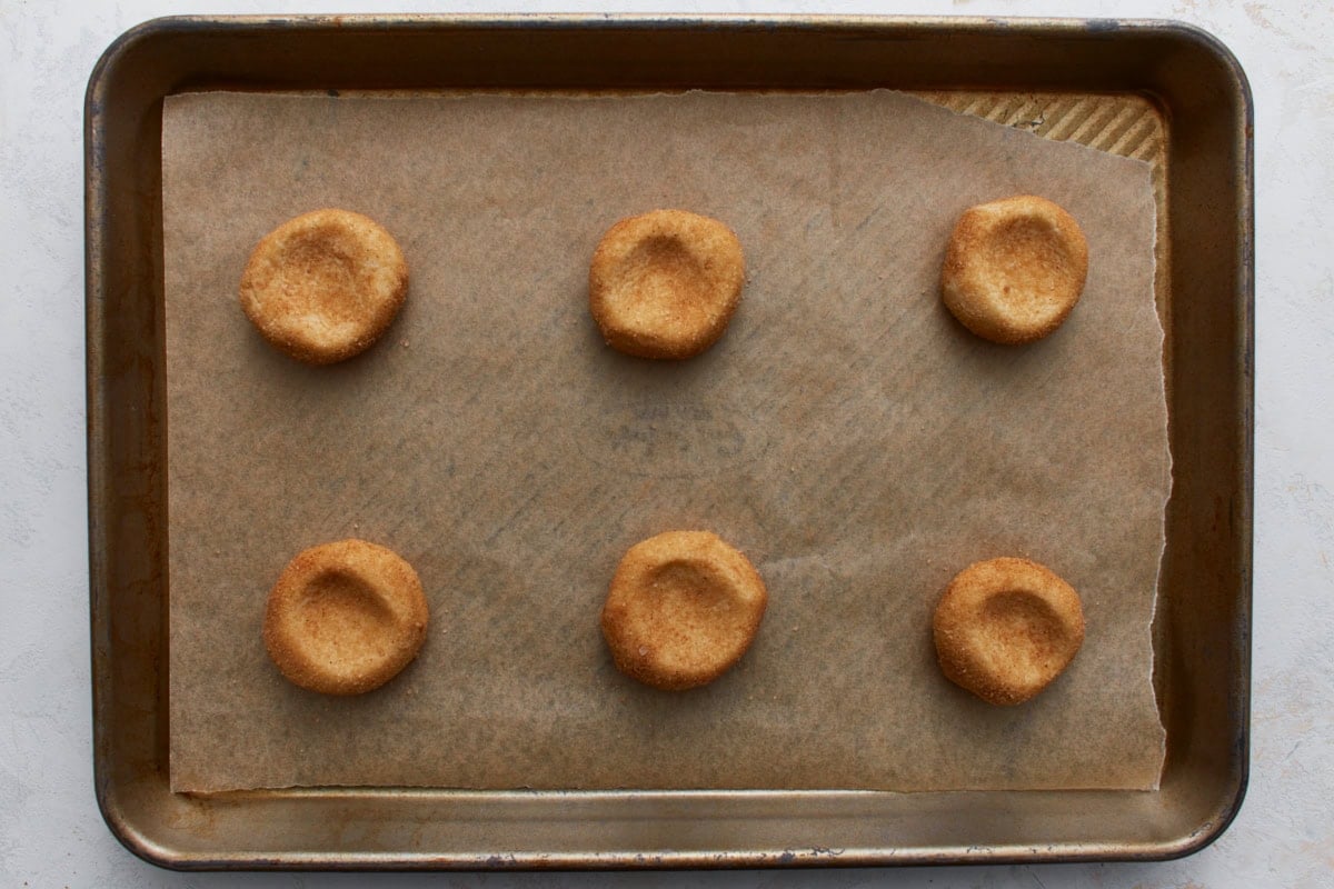 Six coated cookie dough balls placed on a parchment-lined baking sheet, each slightly pressed in the center before baking.