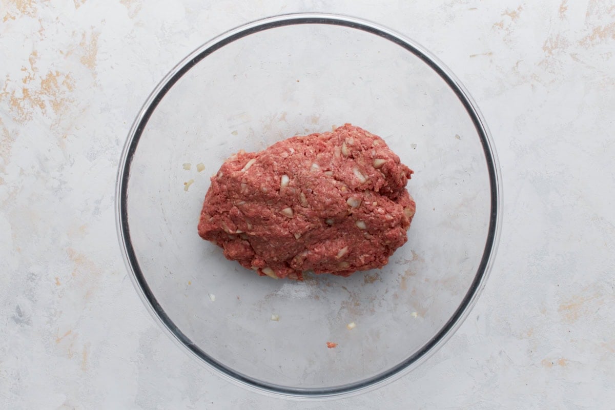 Ground meat mixture with diced onions and seasonings combined in a glass bowl for Swedish meatballs.