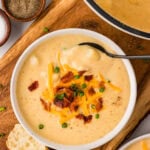 A bowl of creamy gluten-free potato soup topped with shredded cheddar cheese, crispy bacon, and chopped green onions, served with crusty bread on the side.
