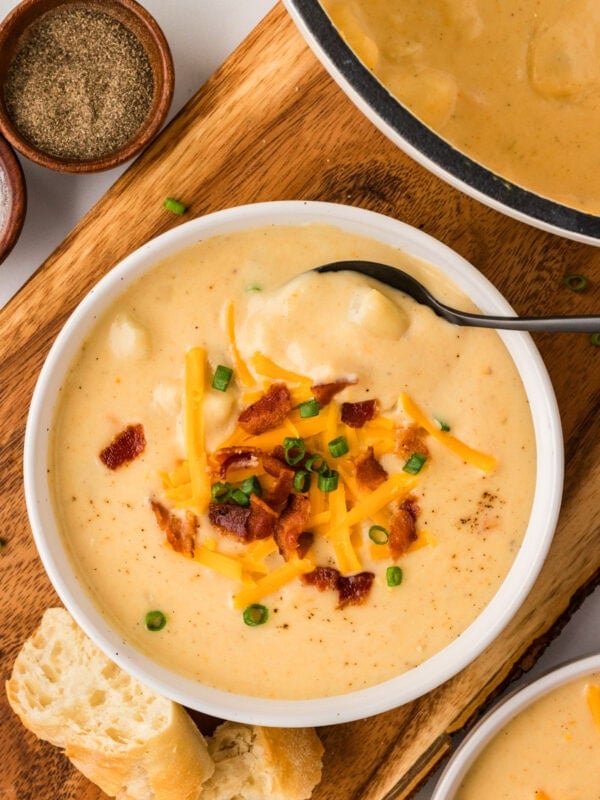 A bowl of creamy gluten-free potato soup topped with shredded cheddar cheese, crispy bacon, and chopped green onions, served with crusty bread on the side.
