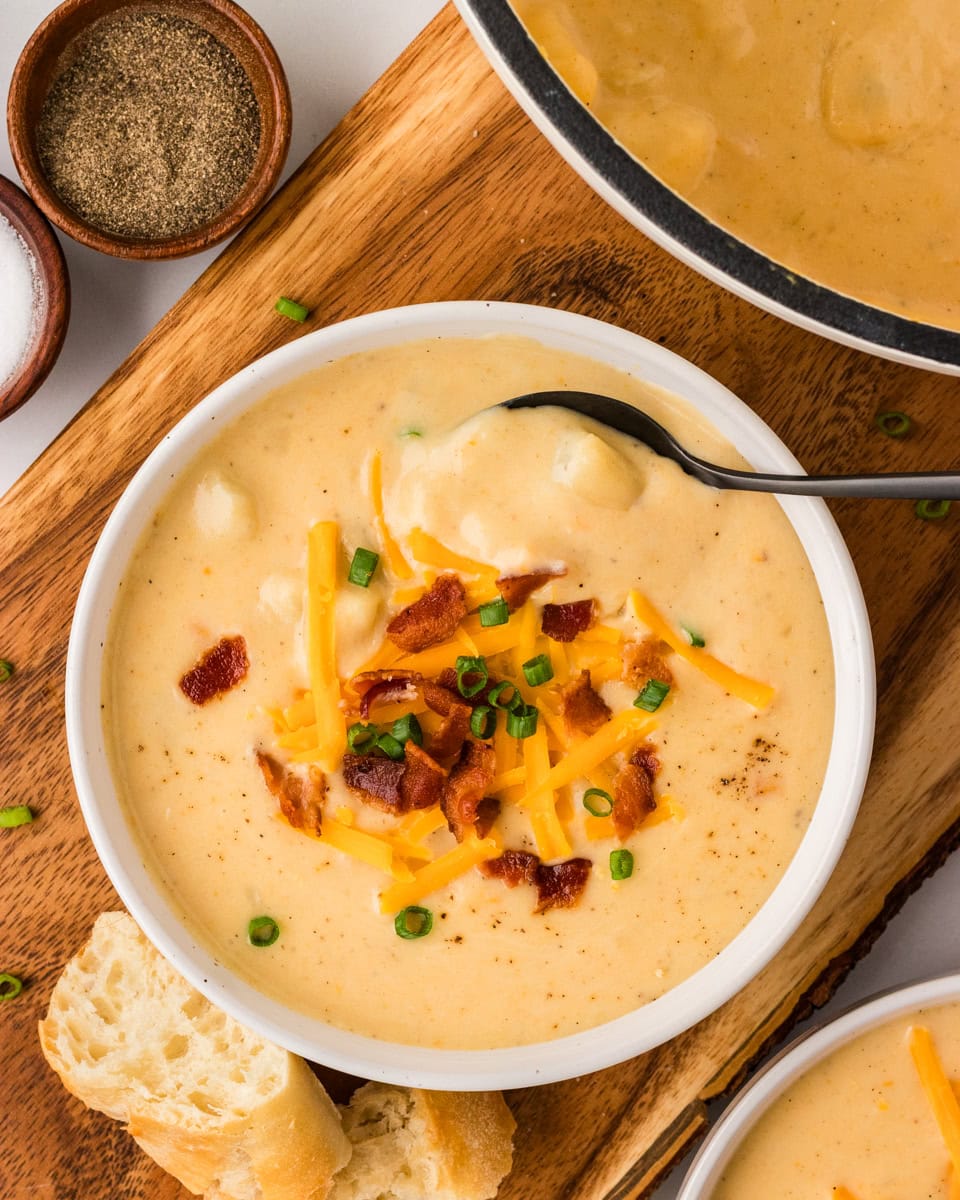 A bowl of creamy gluten-free potato soup topped with shredded cheddar cheese, crispy bacon, and chopped green onions, served with crusty bread on the side.