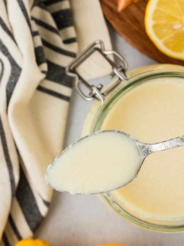 A spoonful of freshly made vegan buttermilk held above a glass jar, showing its creamy texture with lemons and a striped kitchen towel in the background.