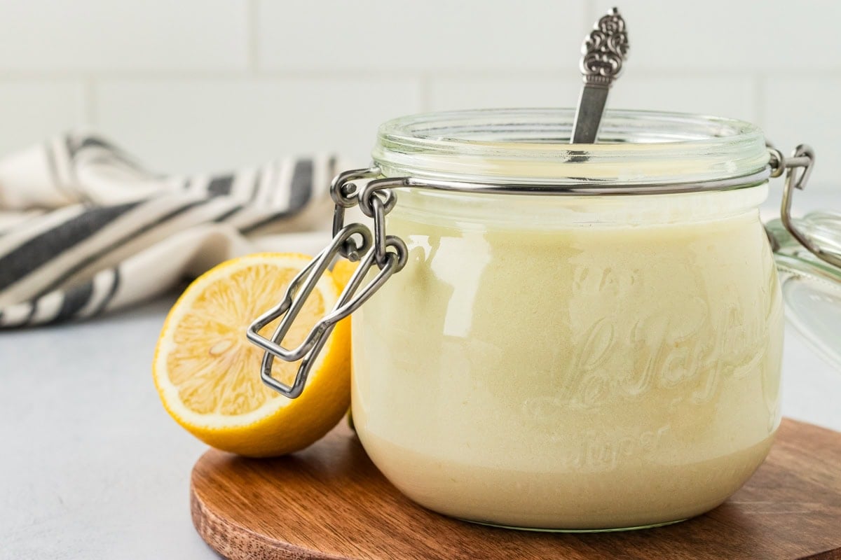 A glass jar filled with creamy vegan buttermilk sits on a wooden board with a spoon inside, next to a halved lemon and a striped kitchen towel in the background.