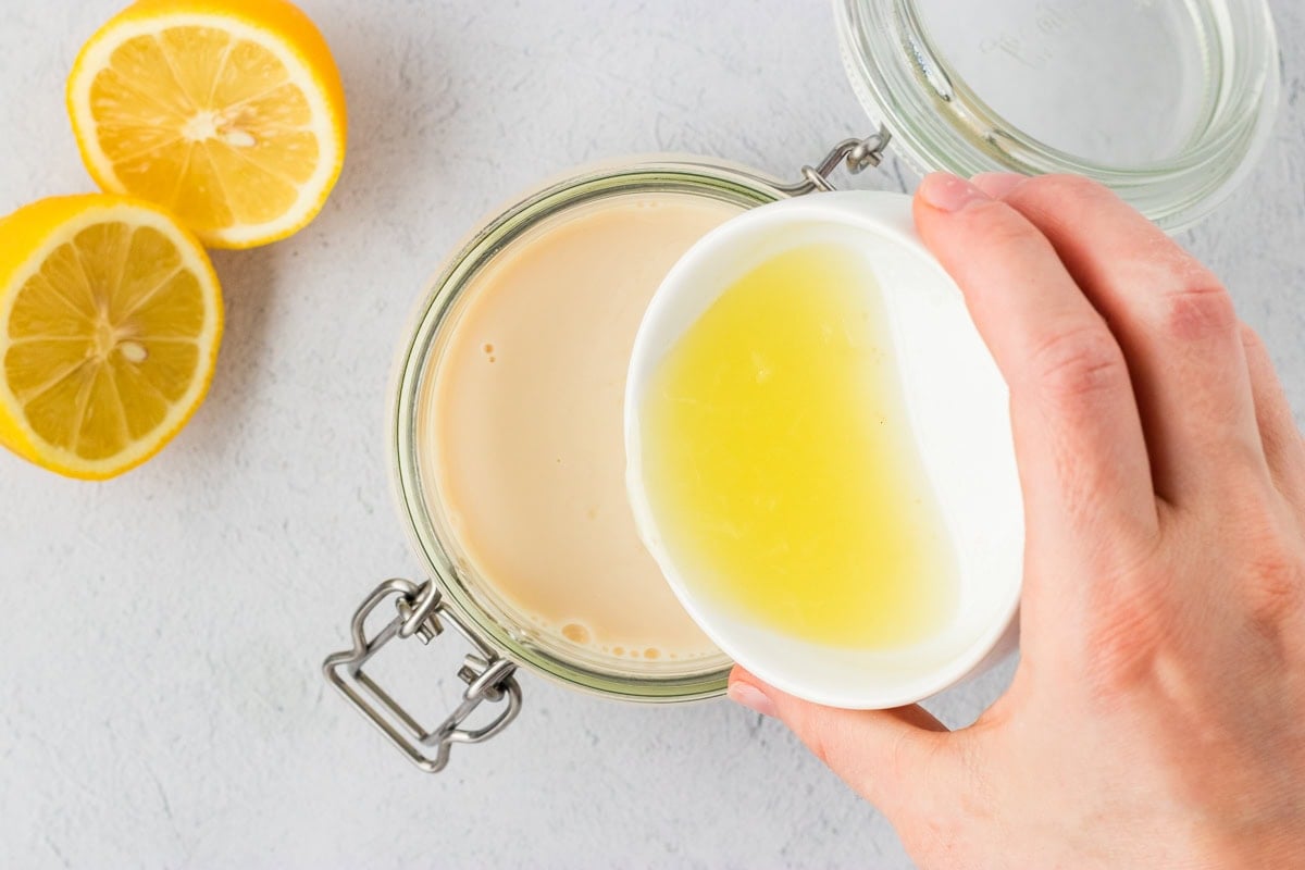 Lemon juice being poured from a small white bowl into a jar of soy milk to make vegan buttermilk, with fresh lemons nearby.
