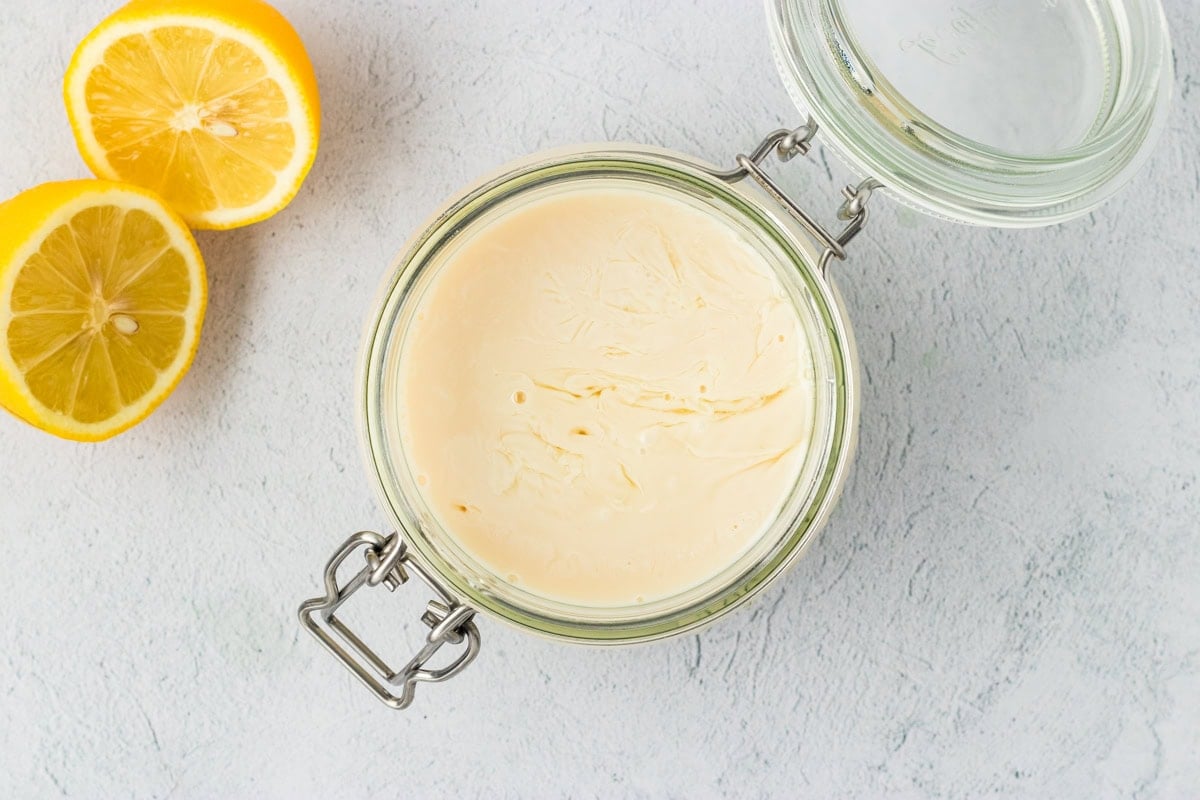 A glass jar filled with freshly curdled vegan buttermilk on a light countertop with halved lemons beside it.