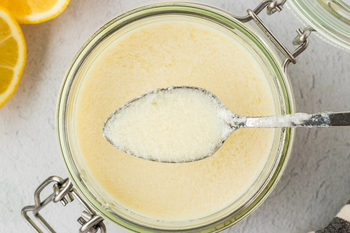 A close-up of a spoon dipped into a jar of vegan buttermilk, showing its lightly curdled, creamy consistency with sliced lemons in the background.