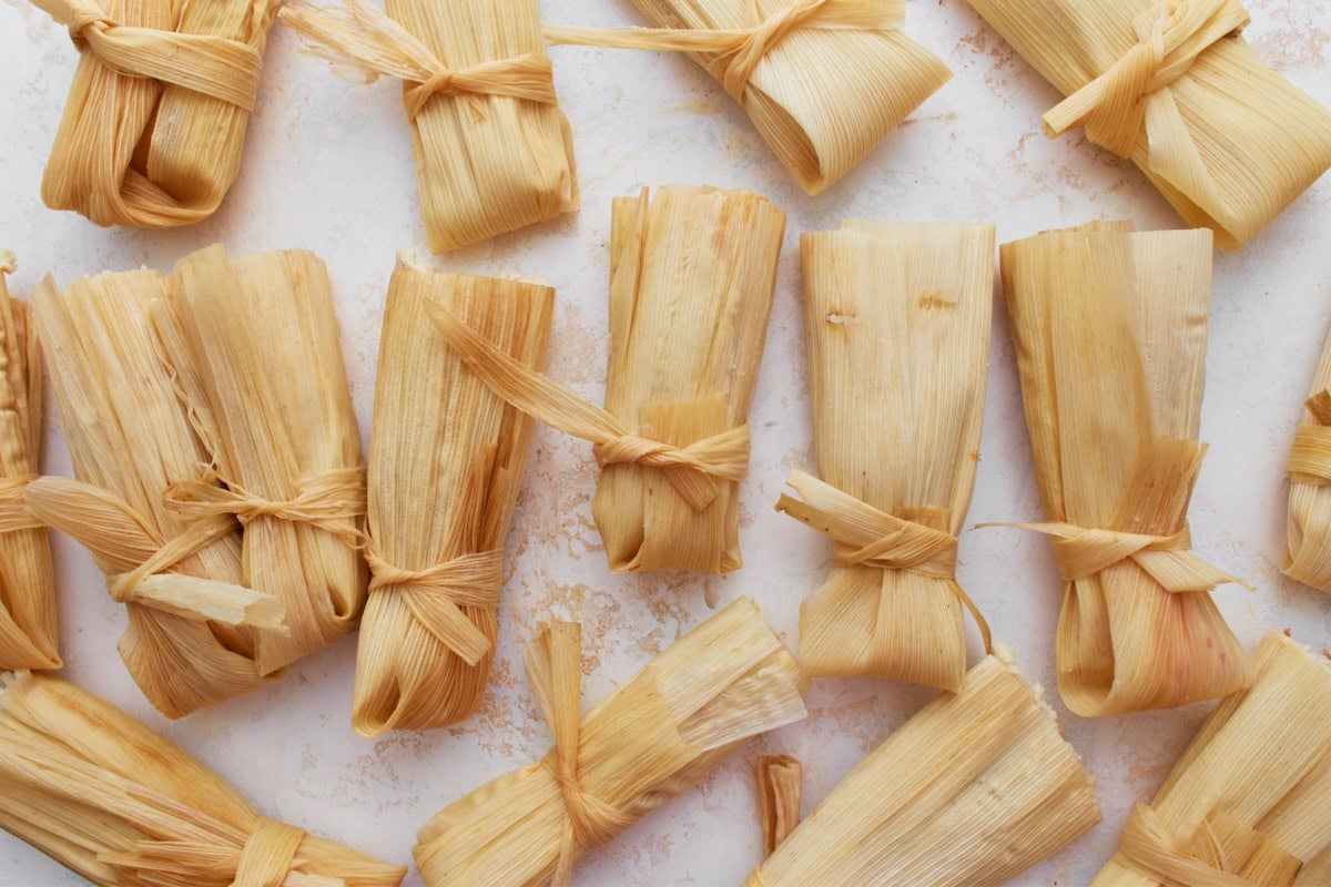 A batch of uncooked tamales wrapped in corn husks and tied with thin strips of husk, ready for steaming.