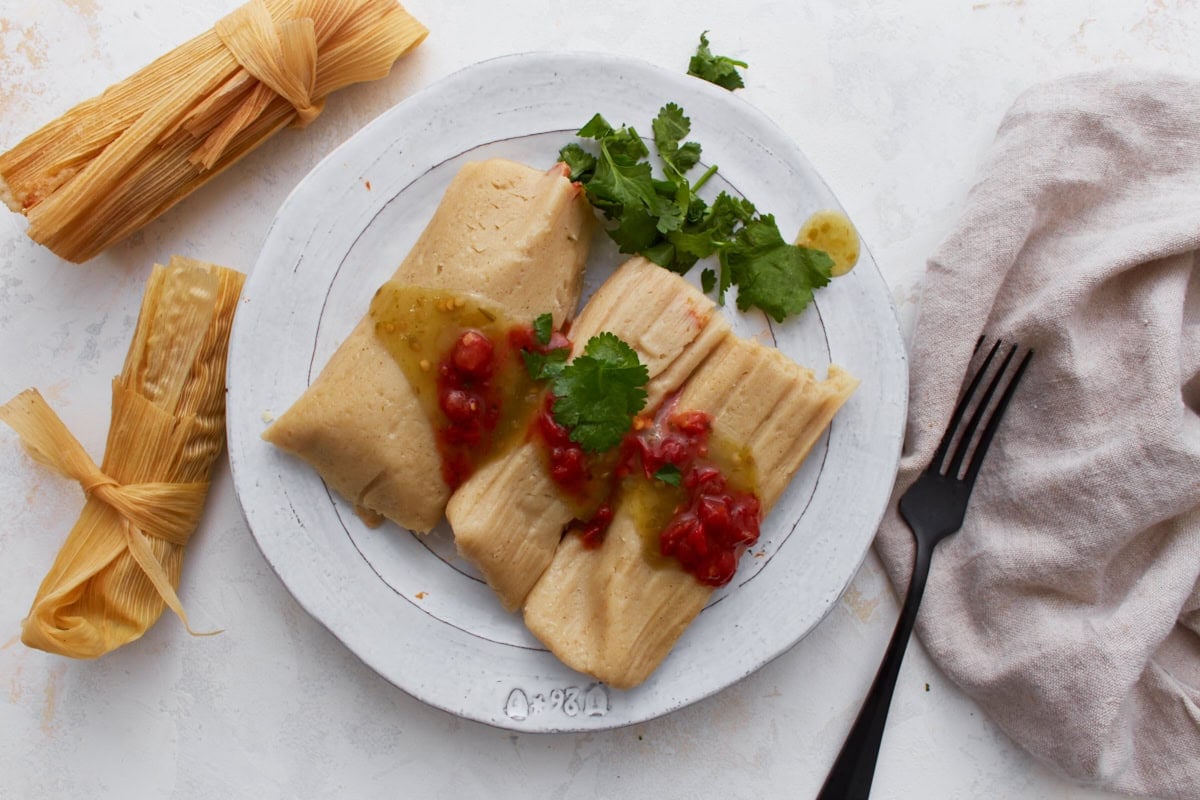 Two gluten-free tamales served on a white plate topped with green salsa and red sauce, garnished with fresh cilantro.