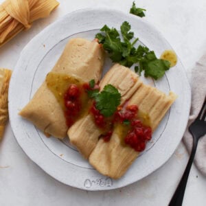 Two gluten-free tamales on a white plate topped with salsa verde and red enchilada sauce, garnished with fresh cilantro.