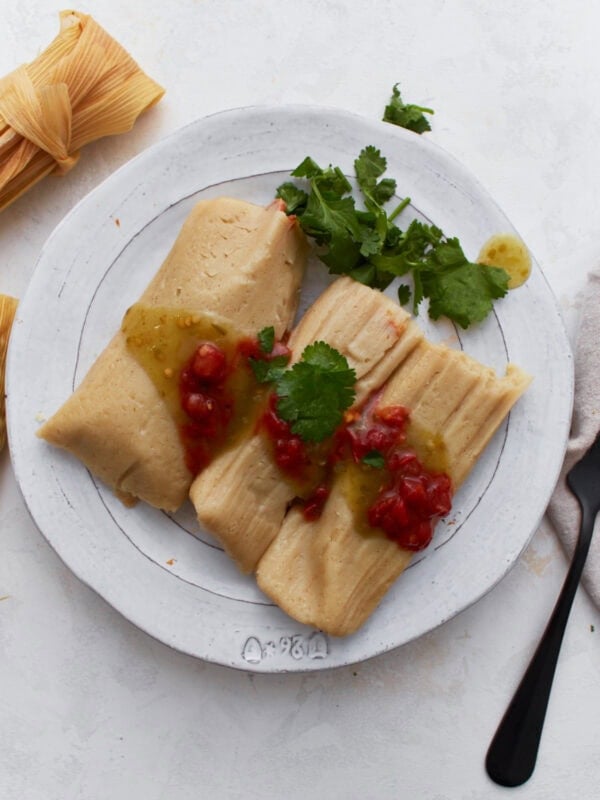 Two gluten-free tamales on a white plate topped with salsa verde and red enchilada sauce, garnished with fresh cilantro.