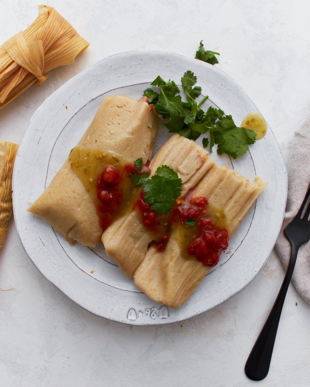 Two gluten-free tamales on a white plate topped with salsa verde and red enchilada sauce, garnished with fresh cilantro.