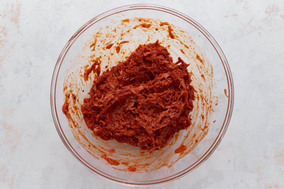 Shredded pork mixed with red enchilada sauce in a glass bowl.