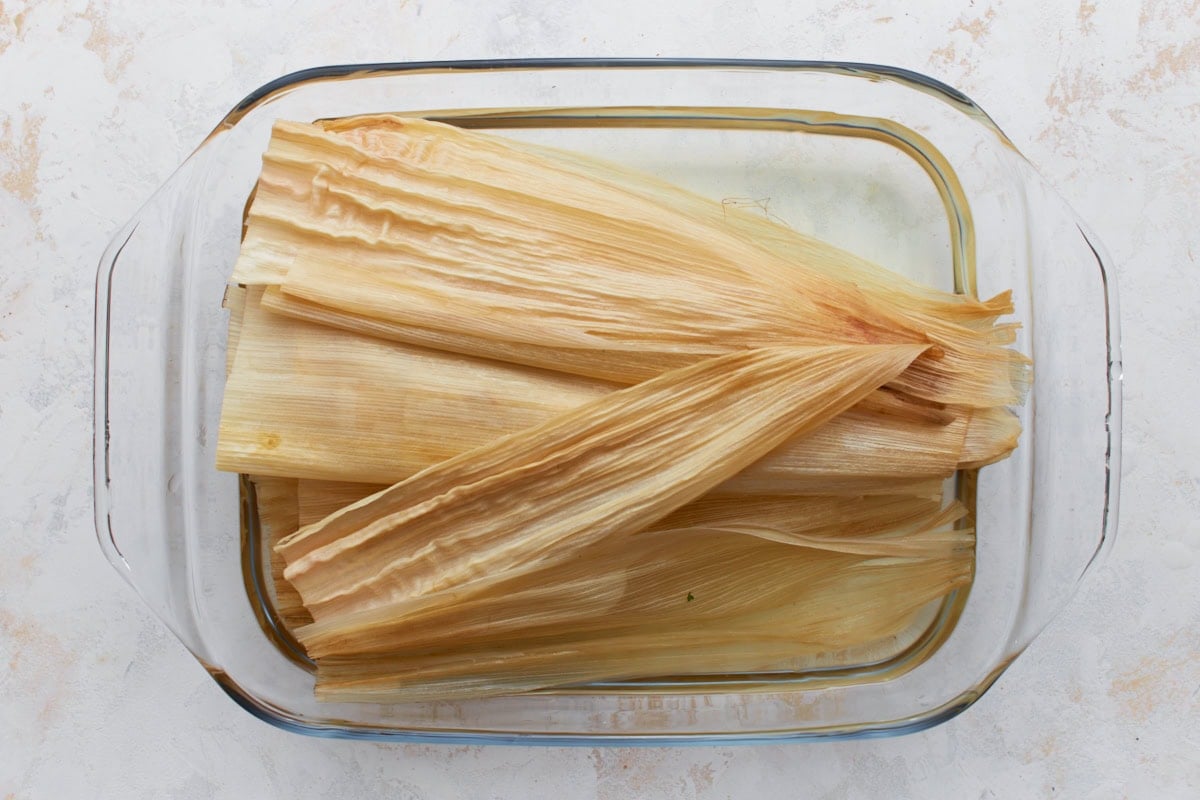 Dried corn husks soaking in hot water inside a glass baking dish until pliable.