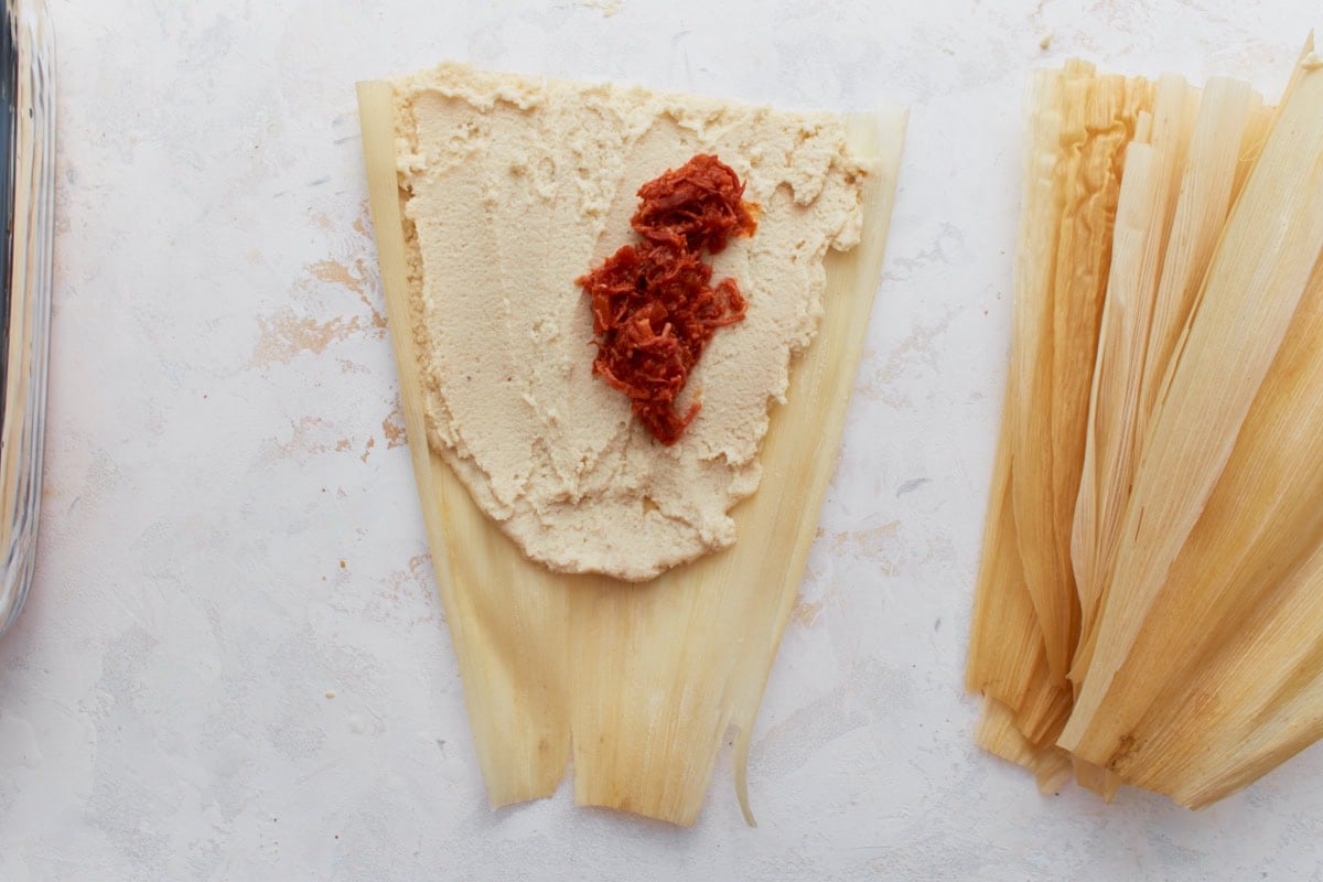 A softened corn husk with masa dough spread over it and a spoonful of pork filling in the center.