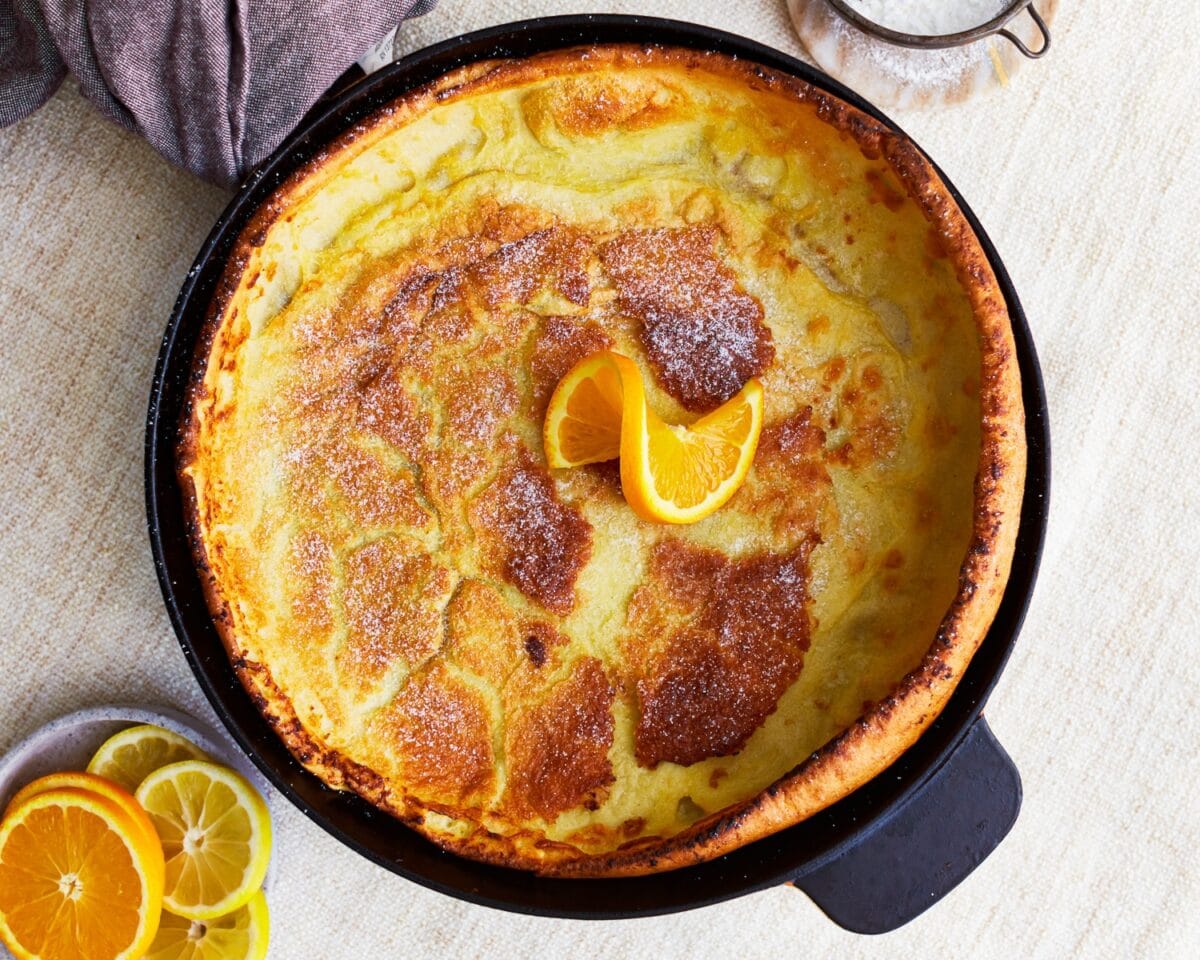 Gluten-free Dutch baby pancake baked golden and dusted with powdered sugar, topped with orange slices and surrounded by lemon and orange wedges.