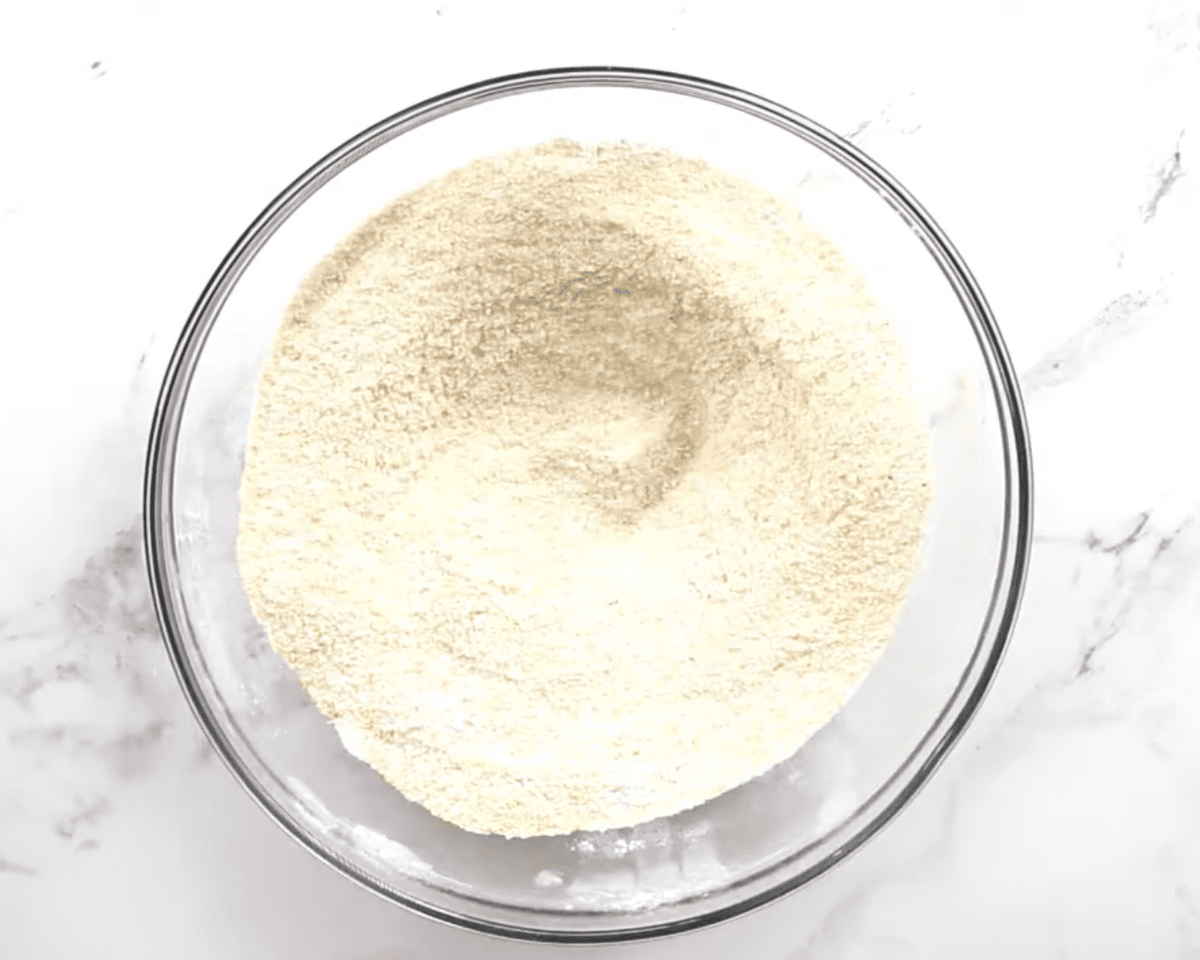 Glass bowl filled with whisked gluten-free cornmeal, flour, and dry ingredients for corn casserole on a white marble surface.