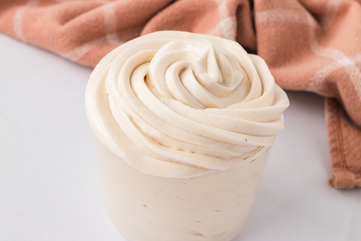 Piping bag-style swirl of dairy-free buttercream frosting in a small container with a soft cloth in the background.