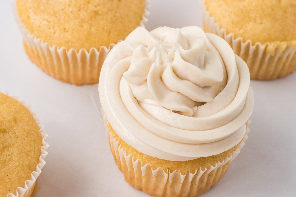 Vanilla cupcake topped with a swirl of dairy-free buttercream frosting surrounded by unfrosted cupcakes.