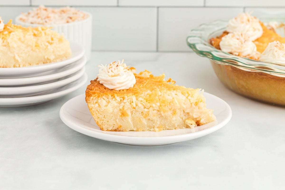 Slice of chilled crustless coconut pie on a white plate topped with whipped cream, with whole pie and plates in the background.