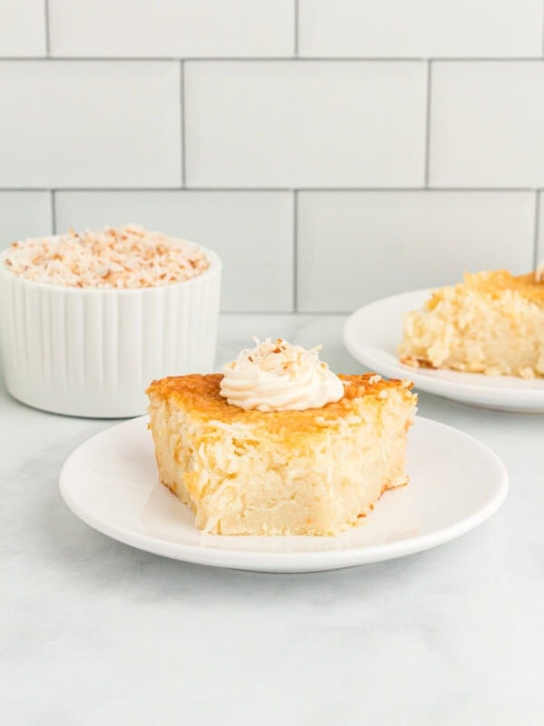Slice of crustless coconut pie on a white plate topped with whipped cream and toasted coconut.