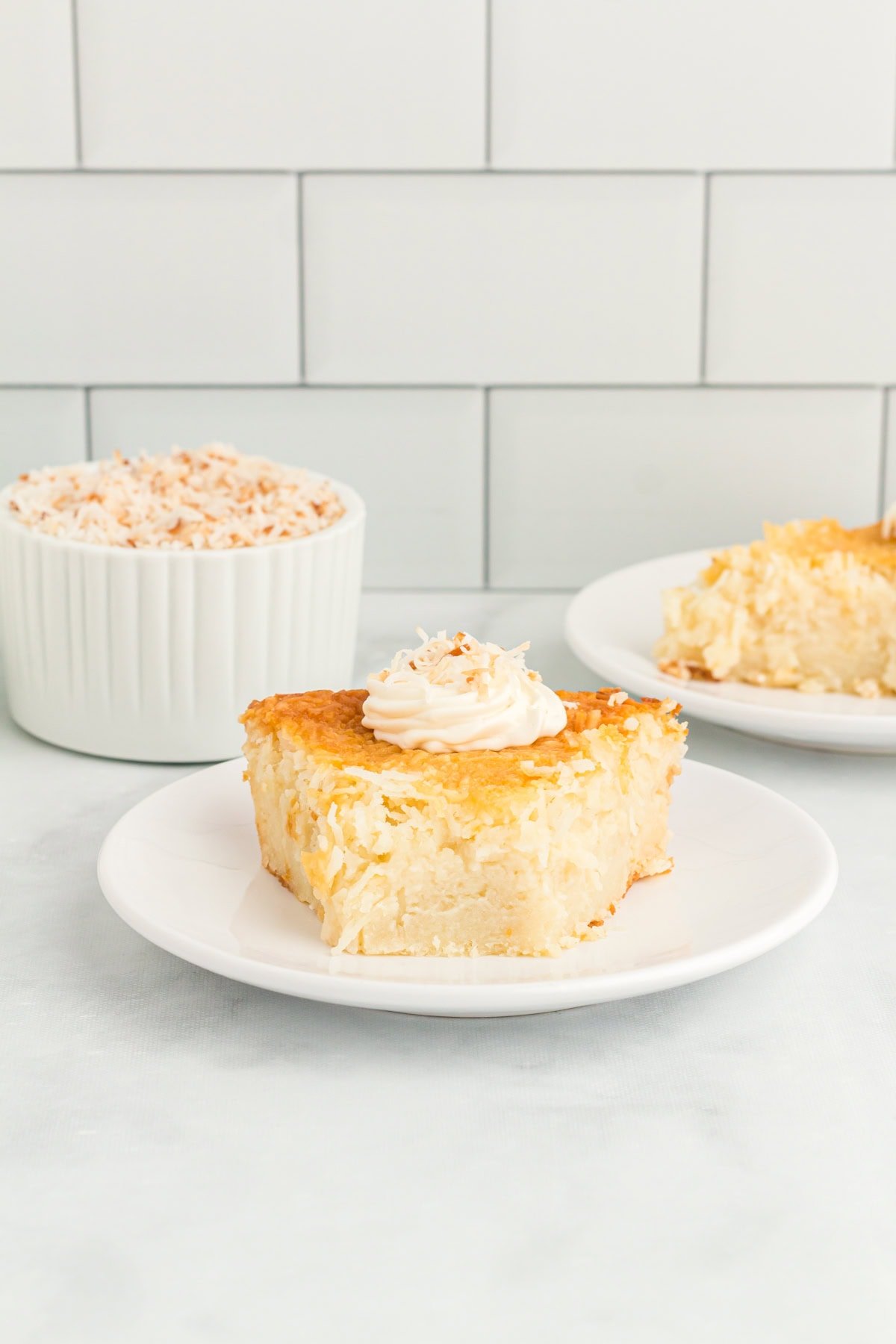 Slice of crustless coconut pie on a white plate topped with whipped cream and toasted coconut.