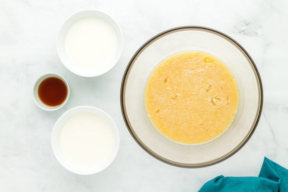 Bowl of whisked sugar, butter, and eggs next to bowls of milk, cream, and vanilla extract.