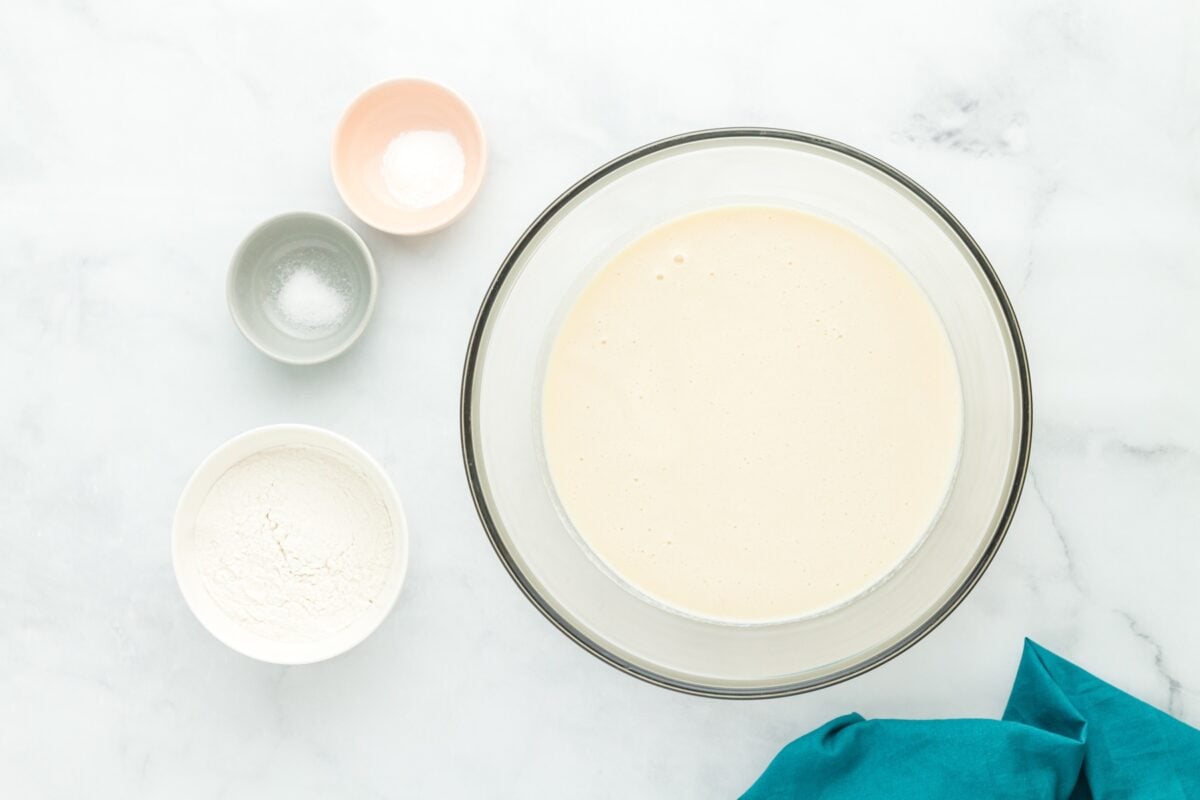 Smooth batter in a glass bowl surrounded by flour, baking powder, and salt in small bowls.