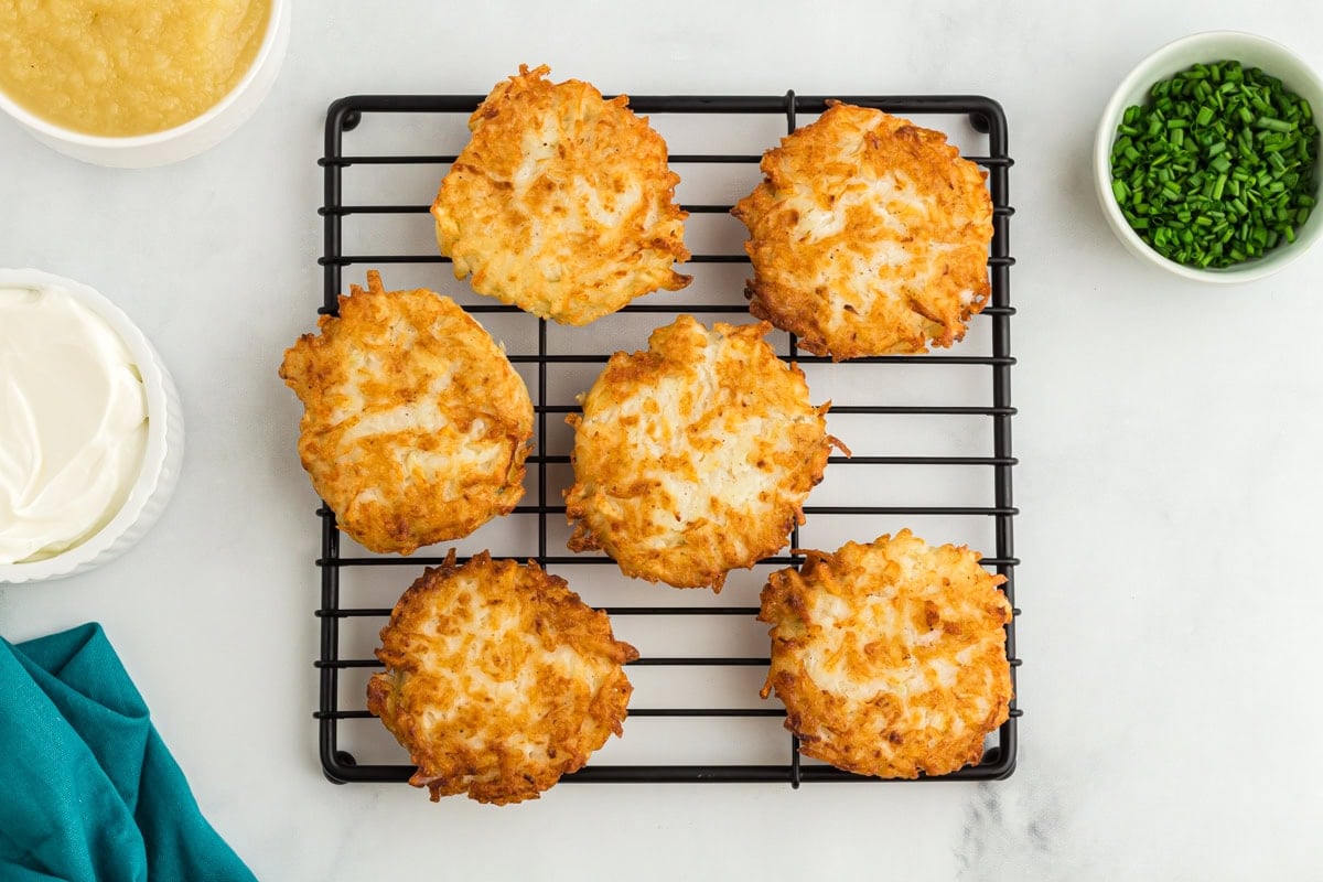 Cooling rack with six crispy gluten-free potato latkes, served alongside bowls of applesauce, sour cream, and chopped chives.