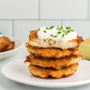 Stack of crispy gluten-free potato latkes topped with sour cream and chopped chives on a white plate.