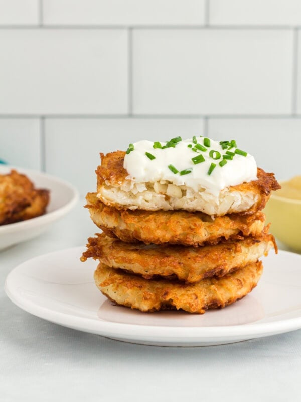 Stack of crispy gluten-free potato latkes topped with sour cream and chopped chives on a white plate.