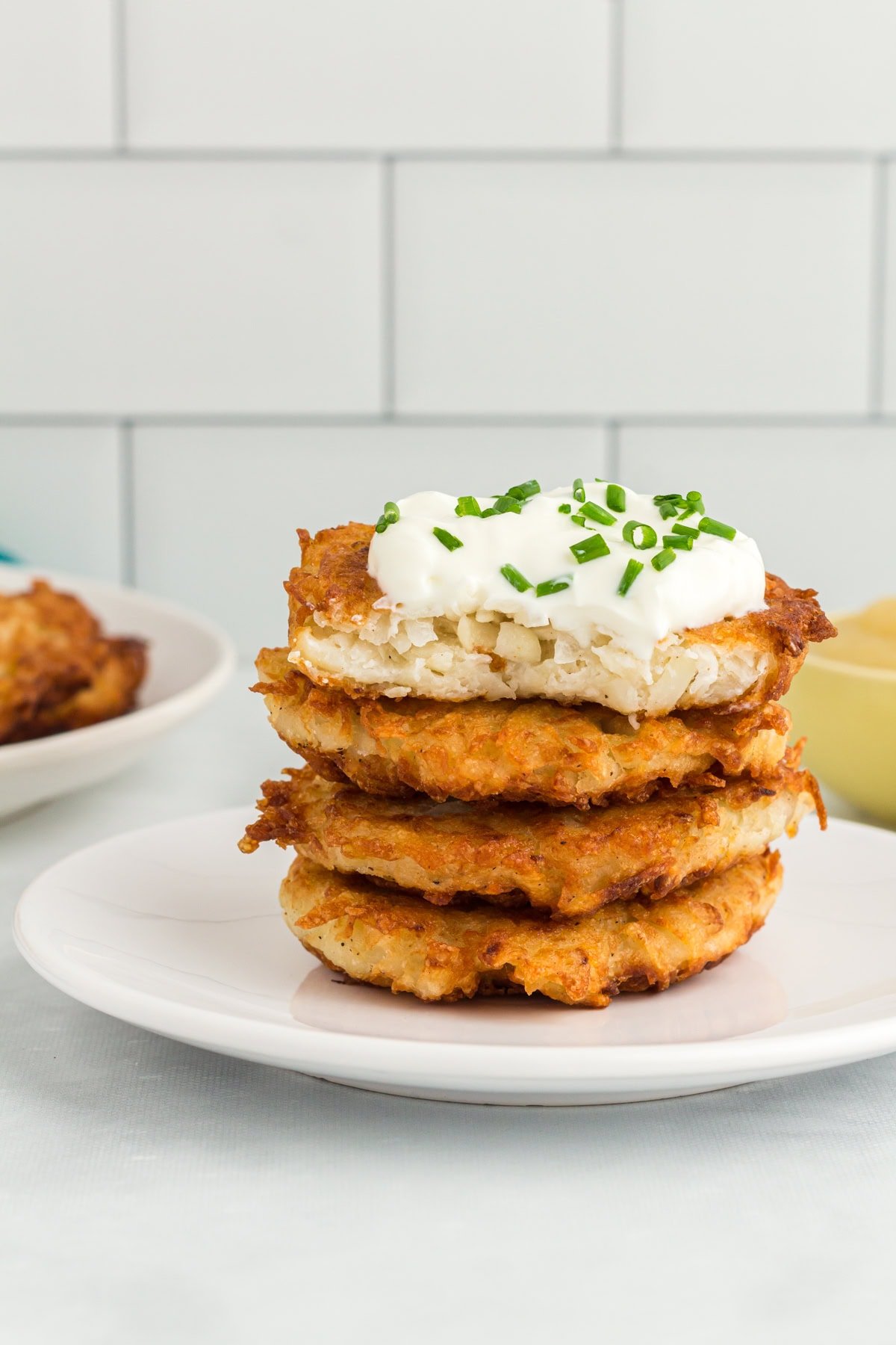 Stack of crispy gluten-free potato latkes topped with sour cream and chopped chives on a white plate.