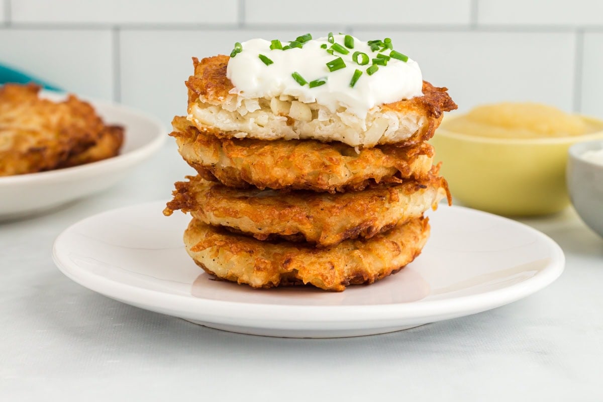 Stack of crispy gluten-free potato latkes topped with sour cream and chopped chives on a white plate.