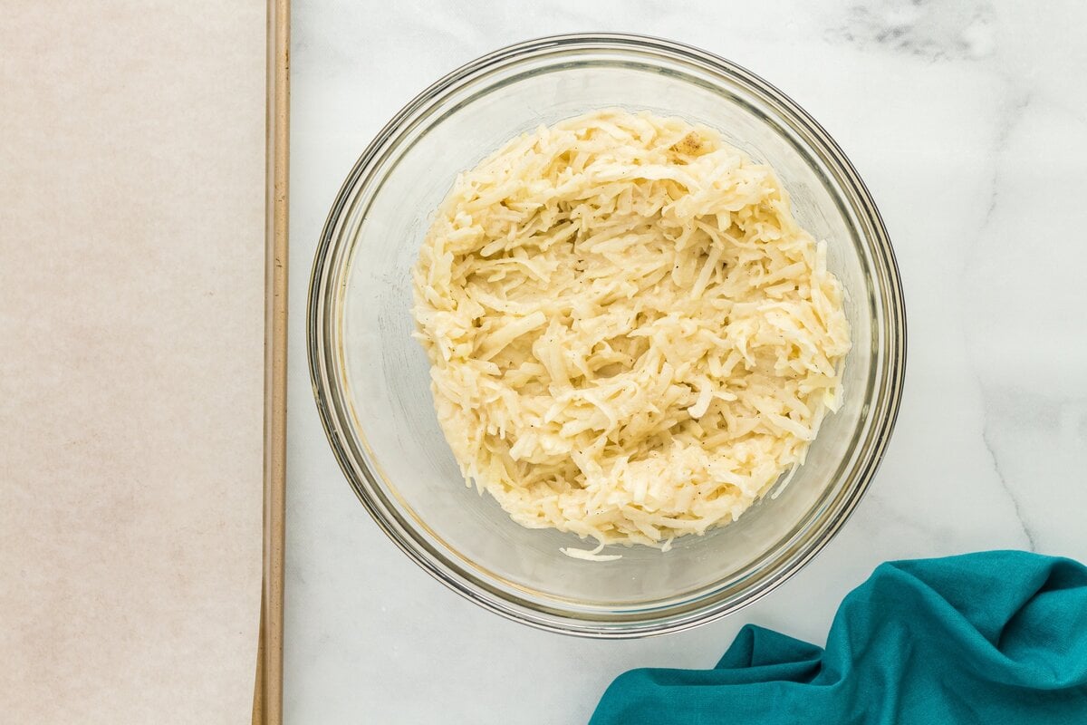 Grated potato and onion mixture combined in a bowl with eggs, gluten-free flour, salt, and pepper.