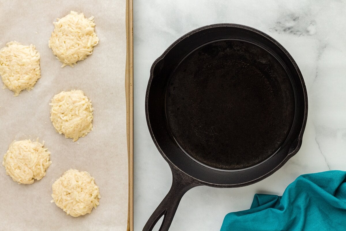 Portioned latke patties shaped on a parchment-lined baking sheet next to a cast-iron skillet.