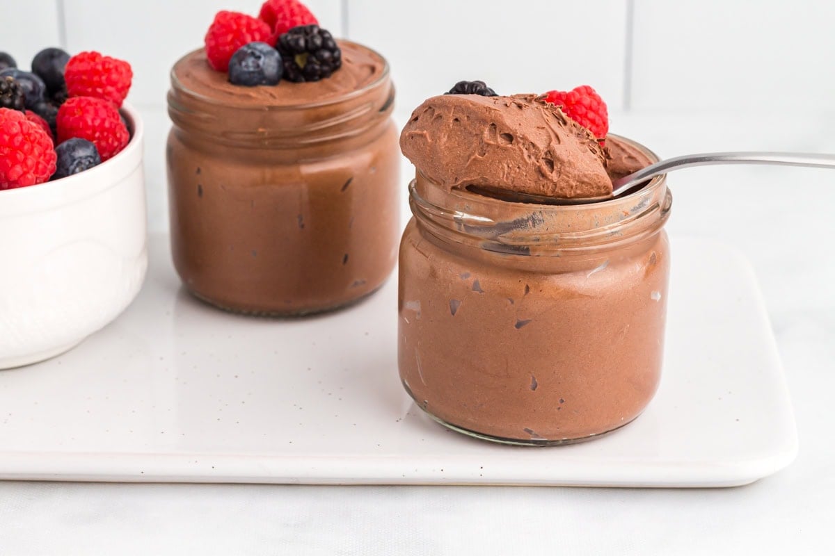 Two jars of vegan chocolate mousse topped with fresh berries, with a spoonful of mousse lifted from one jar and a bowl of mixed berries beside them.